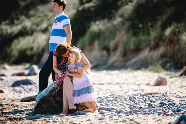 Mama-kuschelt-mit-Tochter-Papa-steht-im-Hintergrund-Shooting-am-Strand-Familienfotografie-Fotograf-Rostock - Familienshooting