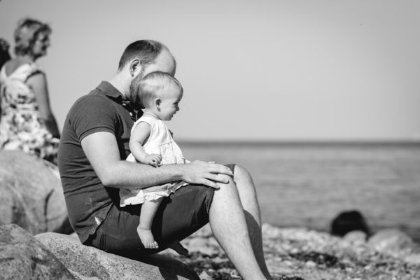 Papa-sitzt-auf-grossem-Stein-mit-seinem-Baby-aud-dem-schoss-Shooting-am-Strand-Familienfotografie-Fotograf-Rostock - Familienshooting