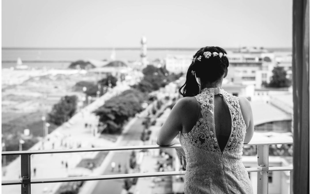 Braut-steht-auf-Balkon-von-Hotel-Neptun-im-Hintergrund-der-Leuchtturm-von-Warnemuende-Hochzeit-am-Strand-Hochzeitsfotograf-Warnemuende-Hochzeitsfotograf-Rostock-Hochzeitsfotograf-Ostseebad-Warnemuende