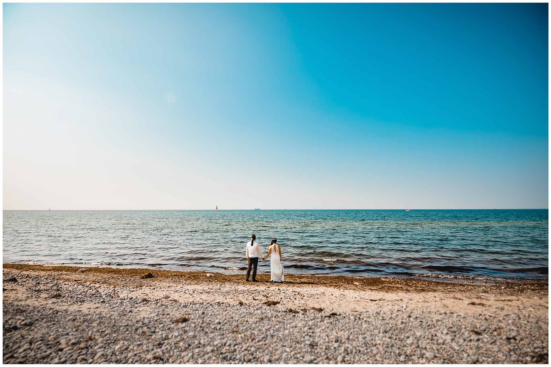 Brautpaar-steht-Hand-in-Hand-direkt-an-der-Ostsee-und-schaut-die-Ferne-Hochzeitsshooting-Hochzeit-am-Strand-Hochzeitsfotograf-Warnemuende-Hochzeitsfotograf-Rostock-Hochzeitsfotograf-Ostseebad-Warnemuende