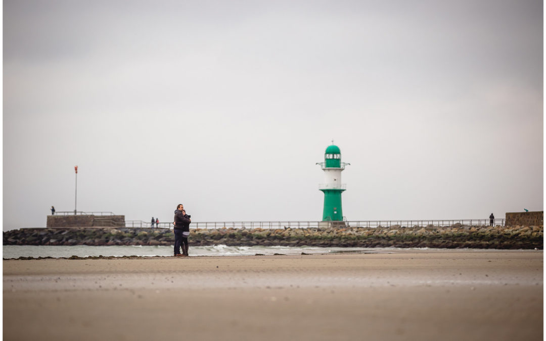 weitwinkelaufnahme-paar-eng-umarmt-steht-am-ostseestrand-im-hintergrund-moewen-und-leuchtturm-mole-warnemuende-shooting-am-strand-fotoshooting-paerchenbilder-fotograf-warnemuende-fotograf-rostock