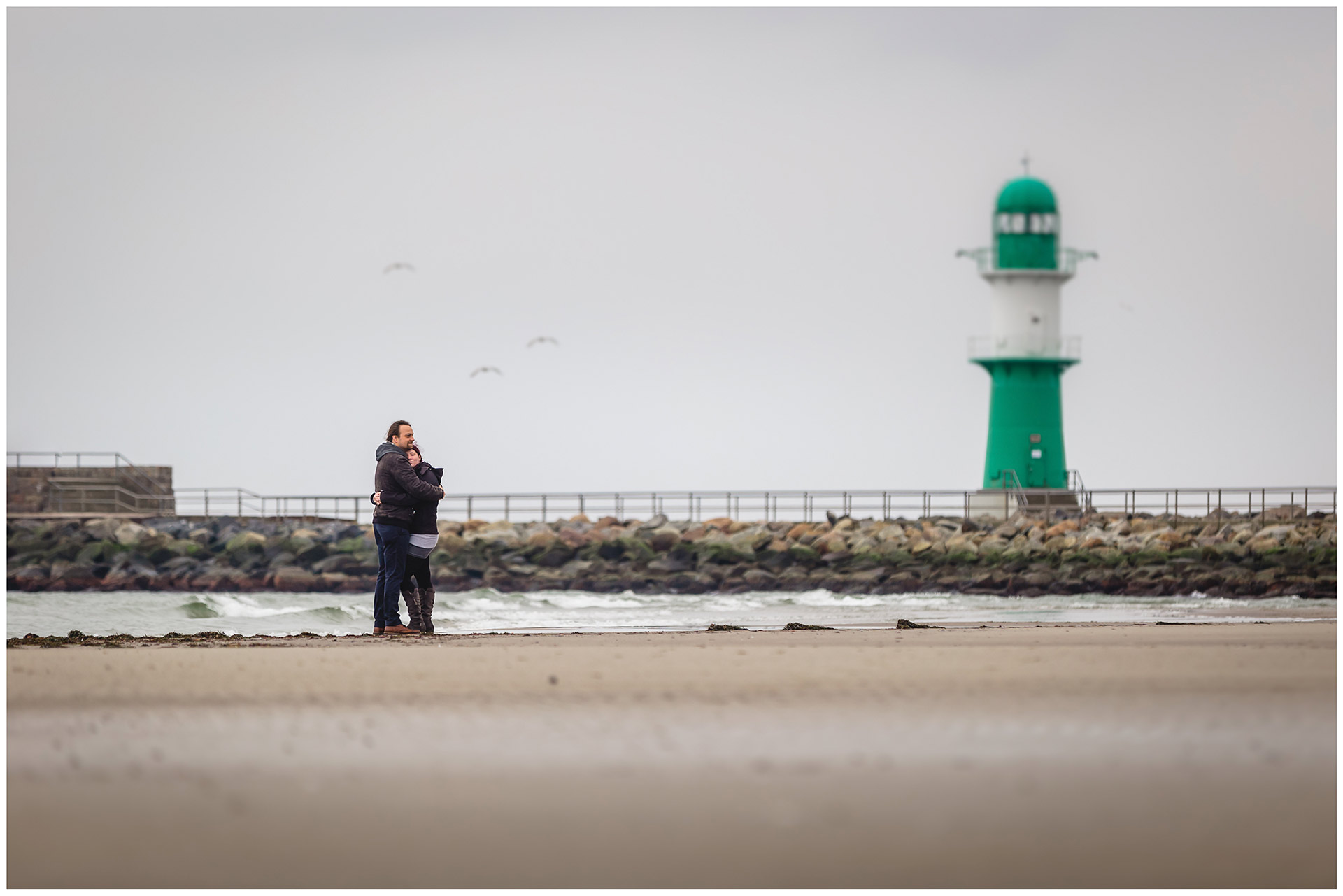 weitwinkelaufnahme-paar-eng-umarmt-steht-am-ostseestrand-im-hintergrund-moewen-und-leuchtturm-mole-warnemuende-shooting-am-strand-fotoshooting-paerchenbilder-fotograf-warnemuende-fotograf-rostock