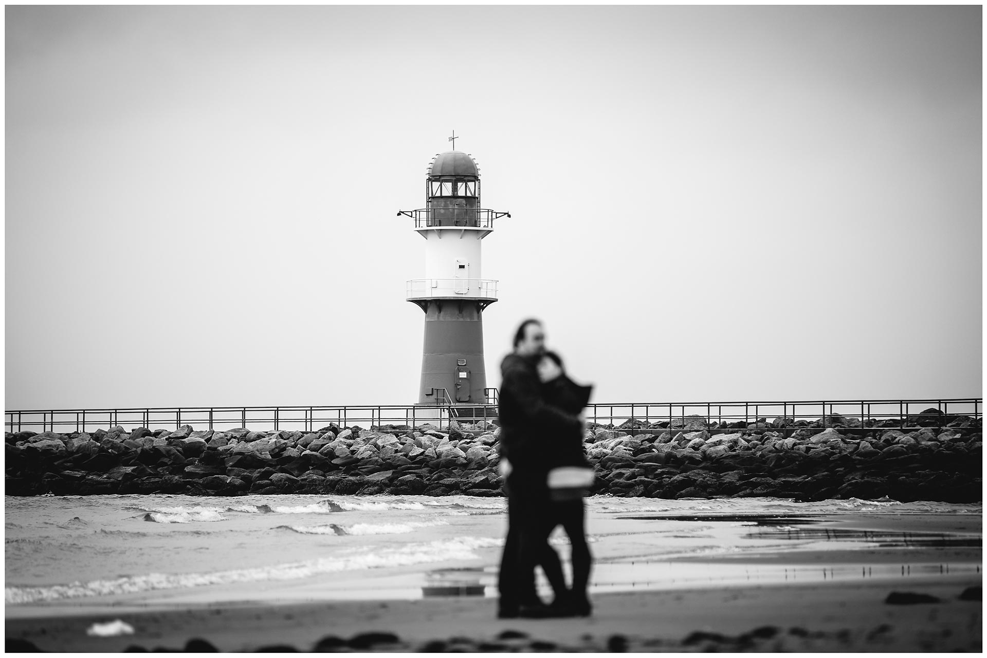 unscharf-im-vordergrund-paar-eng-umarmt-am-ostseestrand-scharf-im-hintergrund-ein-leuchtturm-mole-warnemuende-shooting-am-strand-fotoshooting-paerchenbilder-fotograf-warnemuende-fotograf-rostock