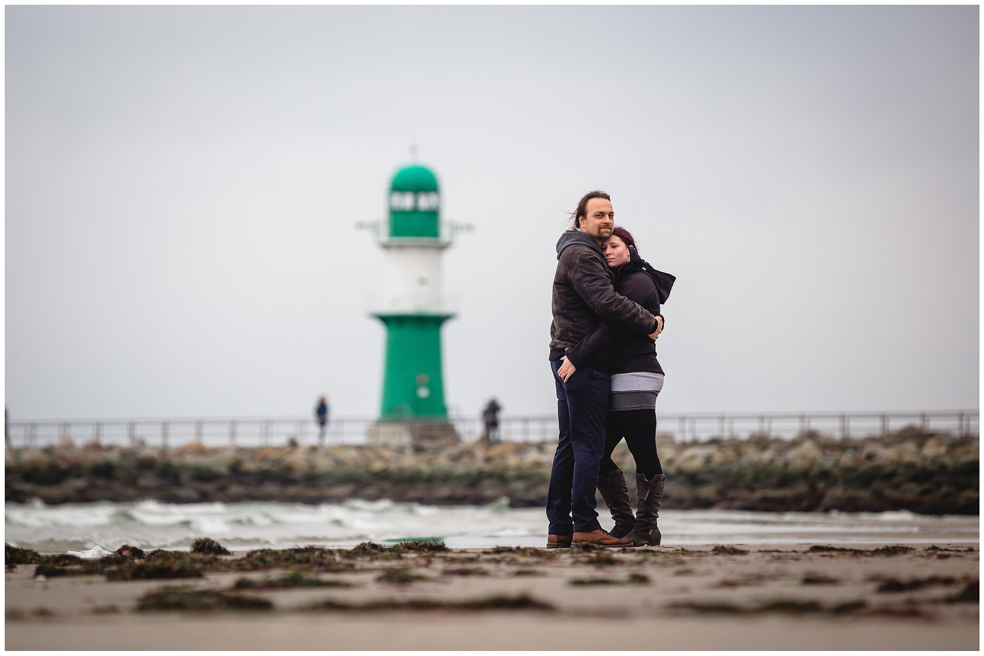 paar-eng-umarmt-steht-am-ostseestrand-im-hintergrund-ein-leuchtturm-mole-warnemuende-shooting-am-strand-fotoshooting-paerchenbilder-fotograf-warnemuende-fotograf-rostock