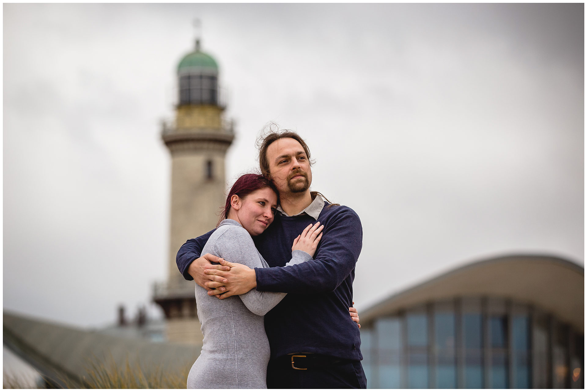 mann-umarmt-frau-ihr-kopf-liegt-an-seiner-schulter-im-hintergrund-teepott-und-leuchtturm-warnemuende-shooting-am-strand-paarshooting-fotograf-rostock-fotograf-warnemünde-paerchenbilder-fotoshooting