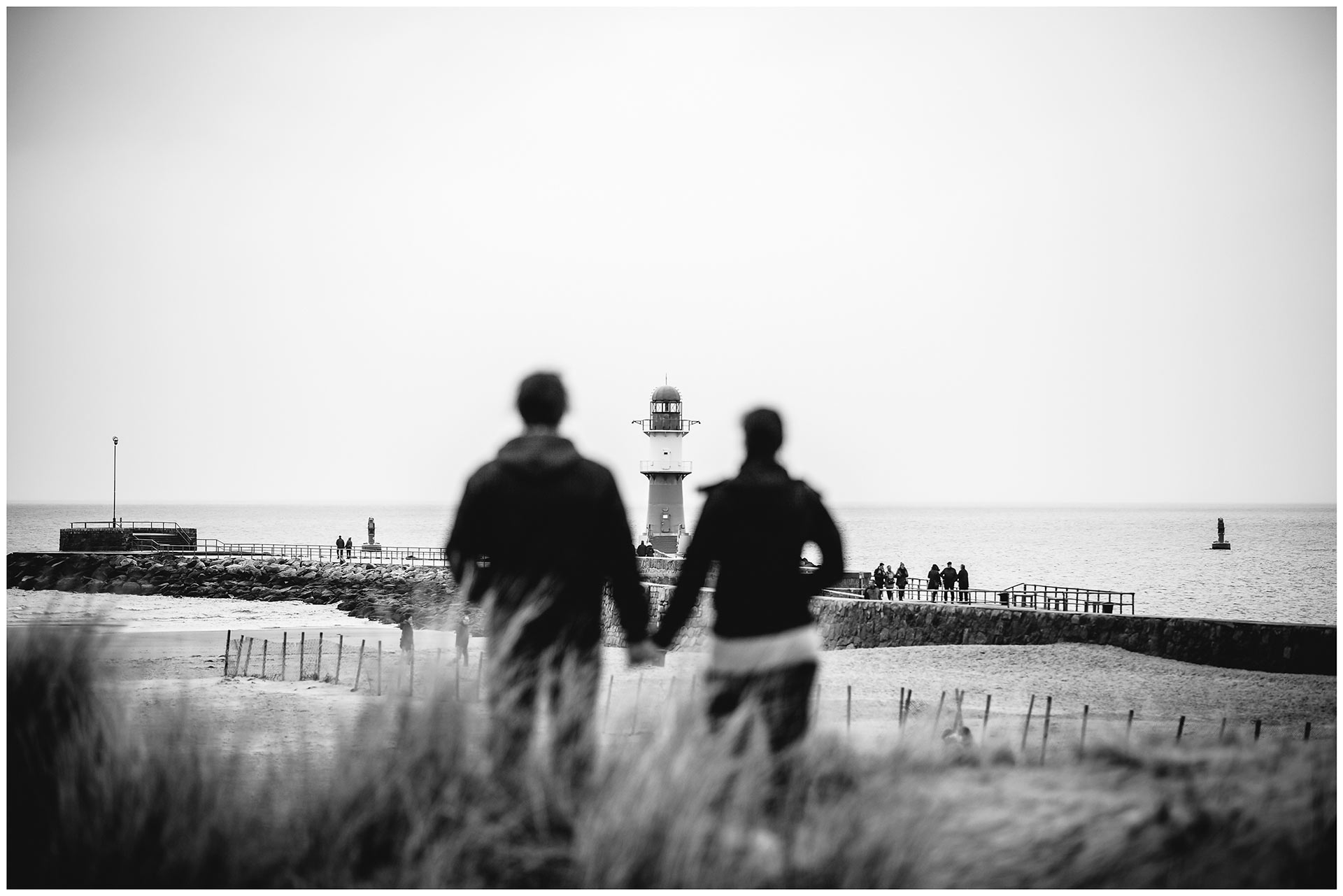 paar-steht-hand-in-hand-auf-duenen-am-strand-schaut-richtung-leuchtturm-warnemuender-mole-shooting-am-strand-paarshooting-fotograf-rostock-fotograf-warnemünde-paerchenbilder-fotoshooting