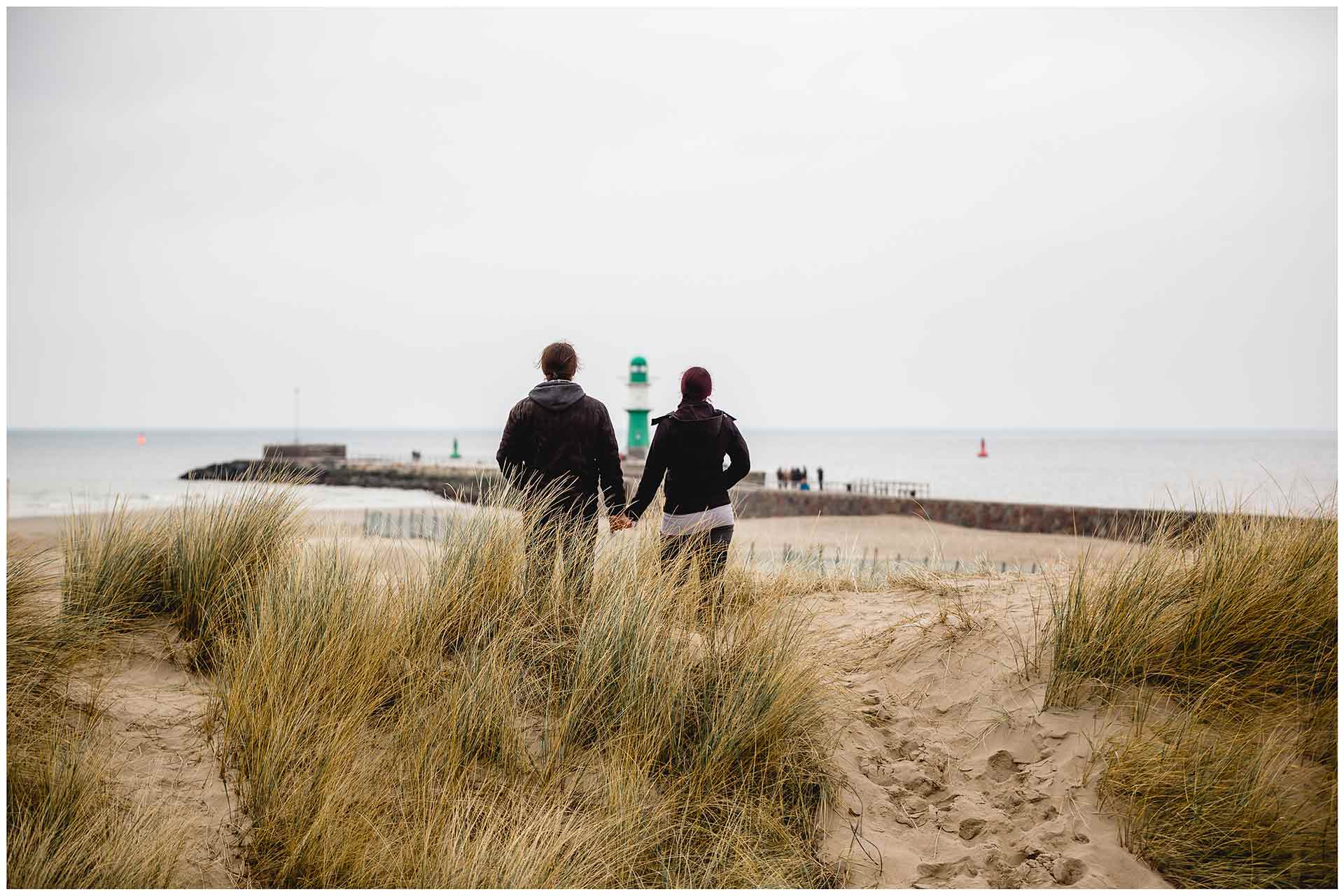 paar-steht-hand-in-hand-auf-duenen-am-strand-schaut-richtung-leuchtturm-warnemuender-mole-shooting-am-strand-paarshooting-fotograf-rostock-fotograf-warnemünde-paerchenbilder-fotoshooting