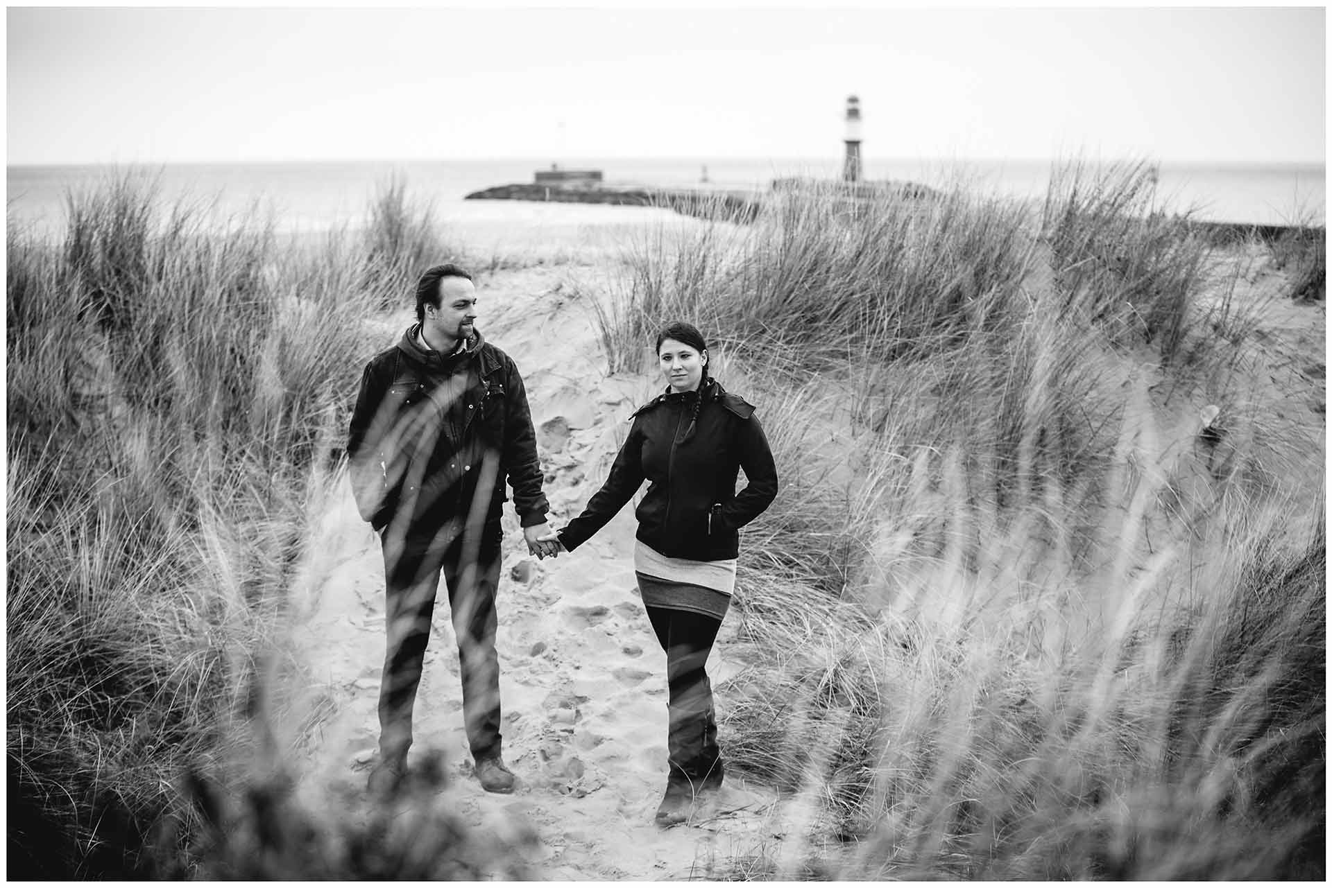 paar-steht-hand-in-hand-auf-duenen-am-strand-schaut-richtung-fotograf-im-hintergrund-leuchtturm-warnemuender-mole-shooting-am-strand-paarshooting-fotograf-rostock-fotograf-warnemünde-paerchenbilder-fotoshooting