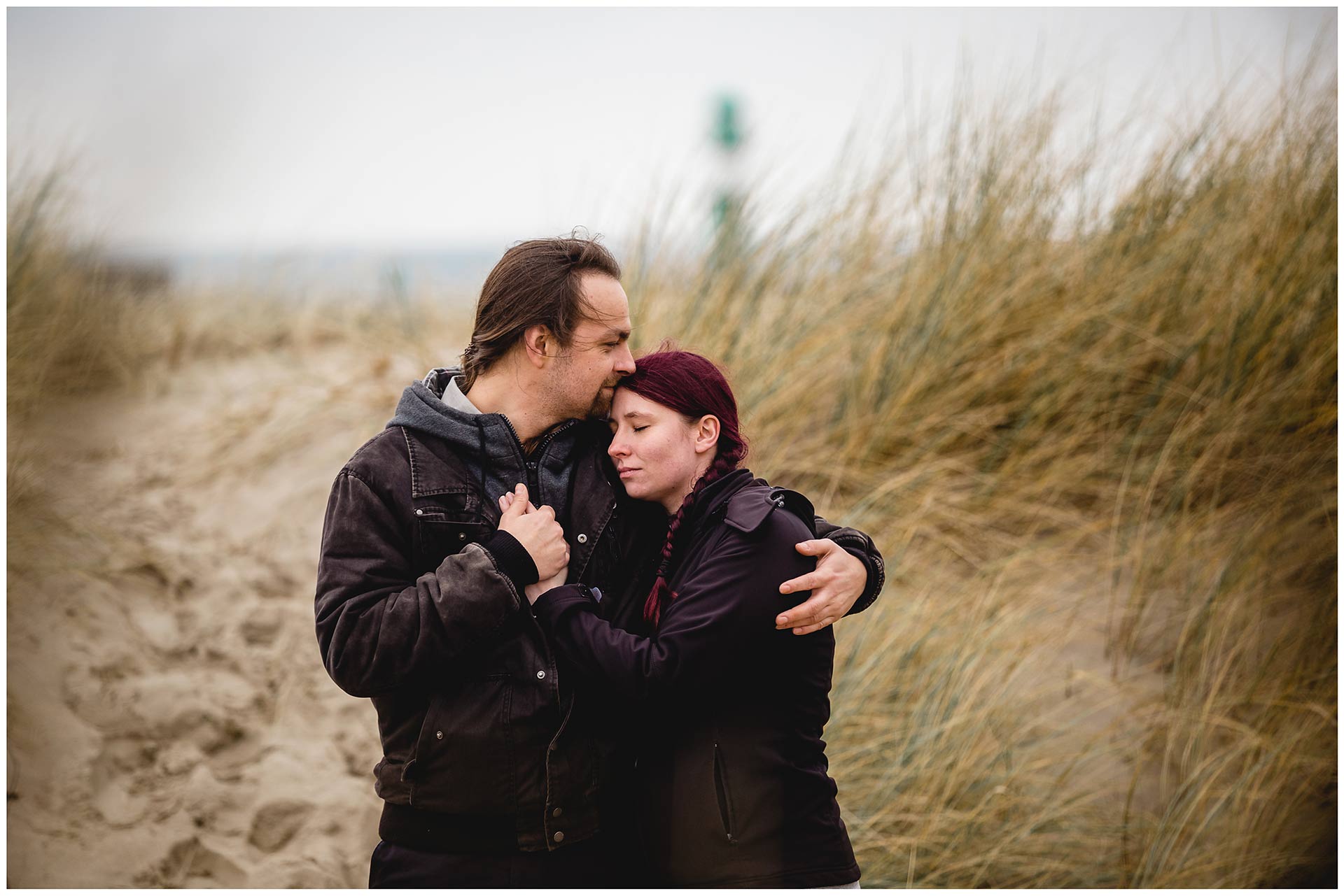 portraitaufnahme-paar-umarmt-sich-eng-und-kuschelt-am-strand-im-hintergrund-leuchtturm-warnemuende-shooting-am-strand-paerchenbilder-fotoshooting-paarshooting-fotograf-rostock-fotograf-warnemünde