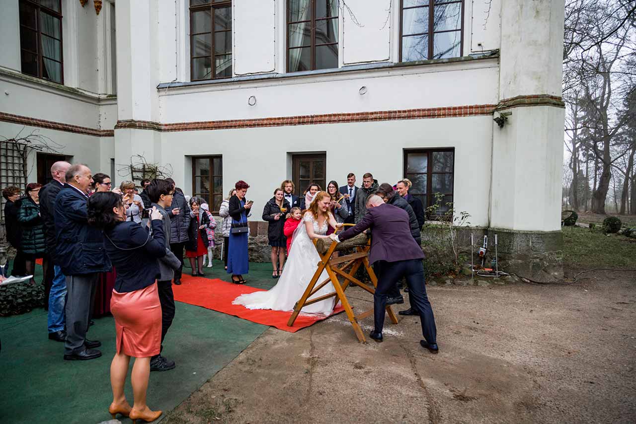 Hochzeitspaar beim Baumstamm sägen im Hintergrund Hochzeitsgäste - Hochzeitsfotograf